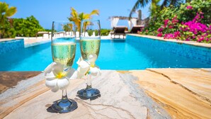 Two champagne glasses with a flower on a stone table by a pool with a clear blue sky and tropical plants in the background. - Next Paradise Boutique Resort (Pwani Mchangani)