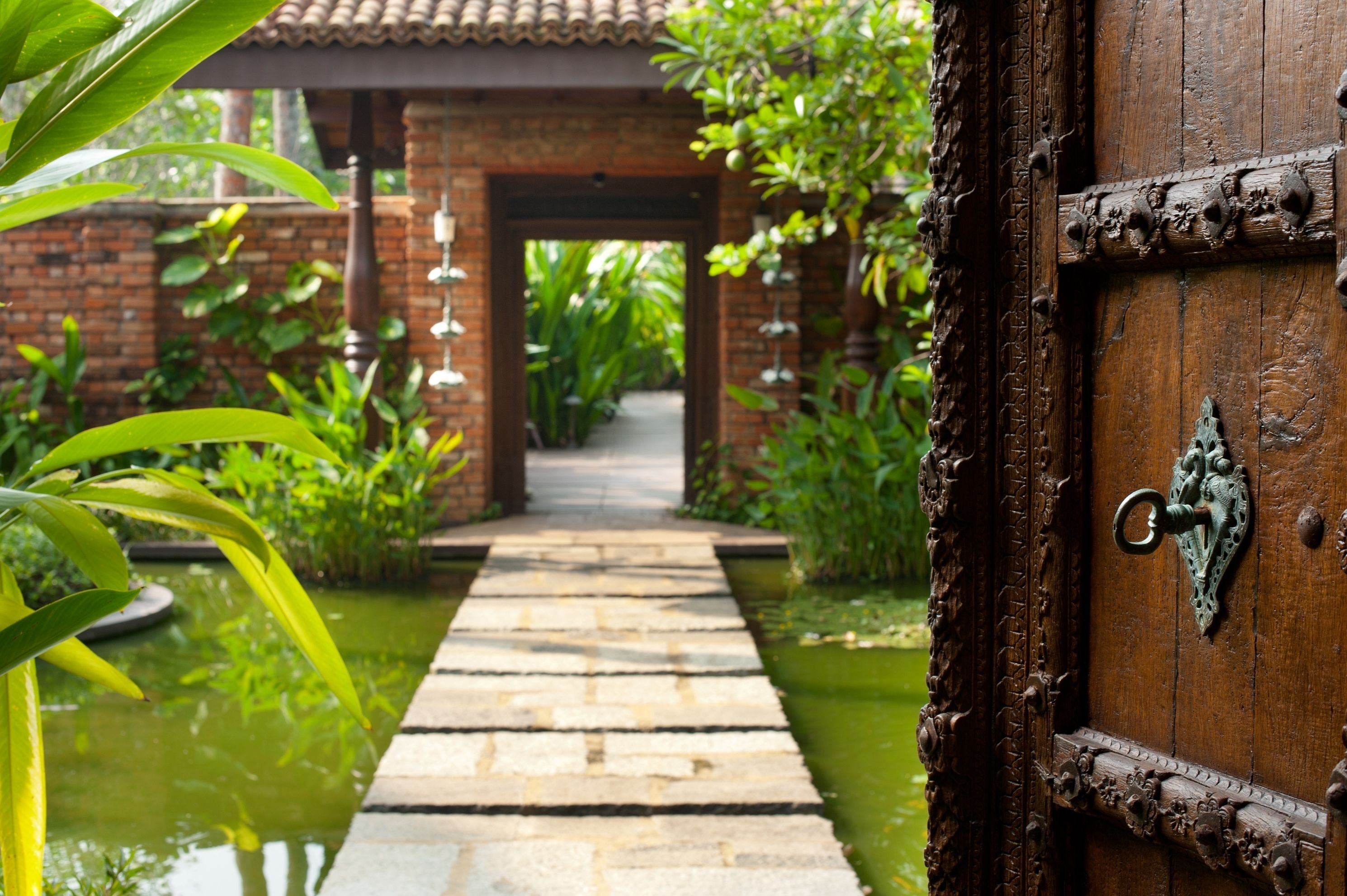 A wooden door with a metal handle, leading to a brick pathway surrounded by greenery.