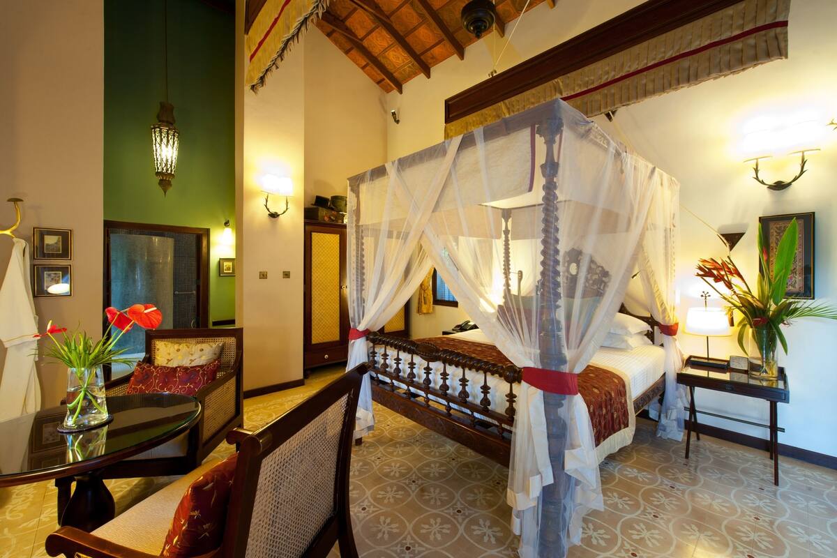 A room with a four-poster bed, a sitting area with a sofa and armchair, a dining table, and a small side table with a lamp.