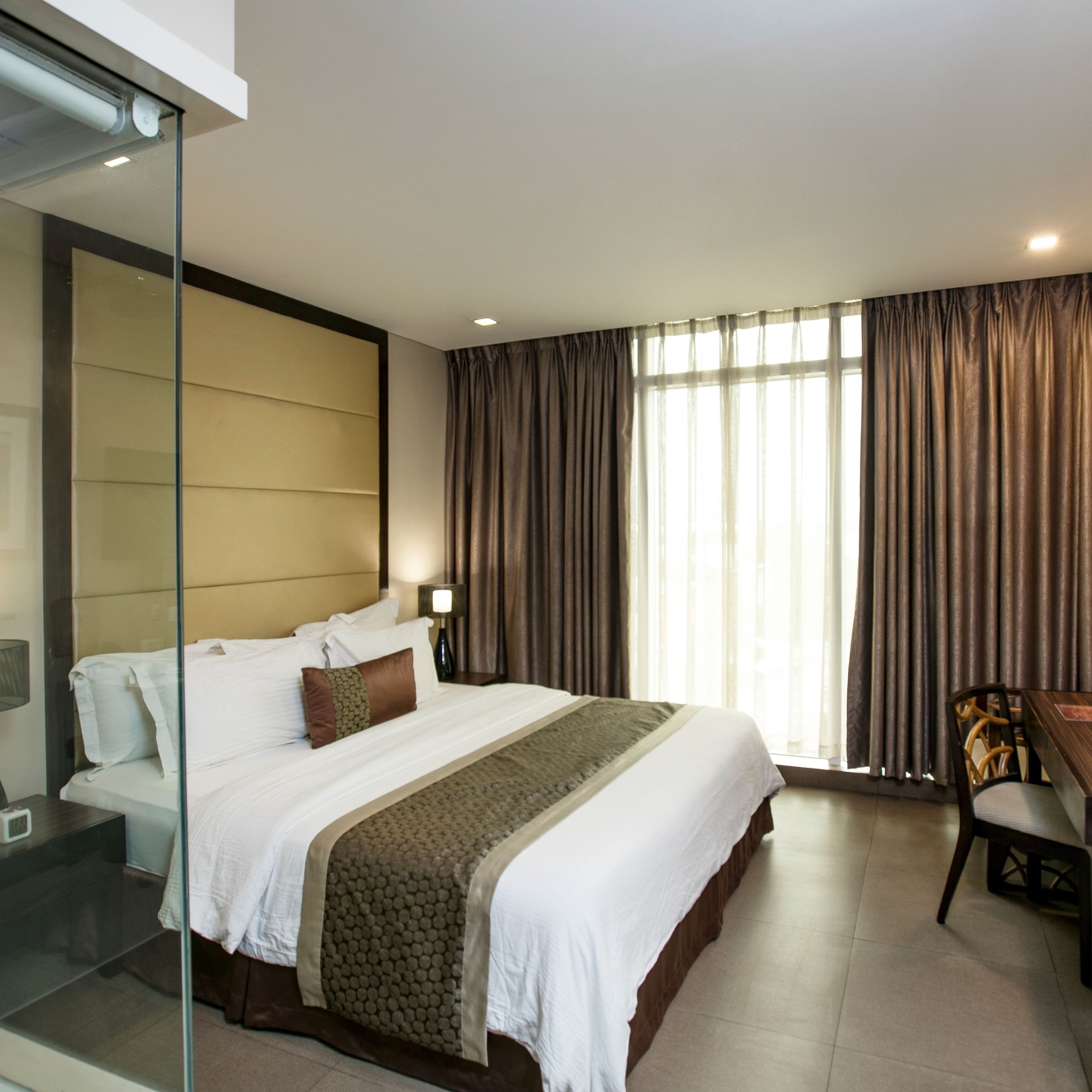 A modern hotel room with a large bed, a desk, and a glass partition.