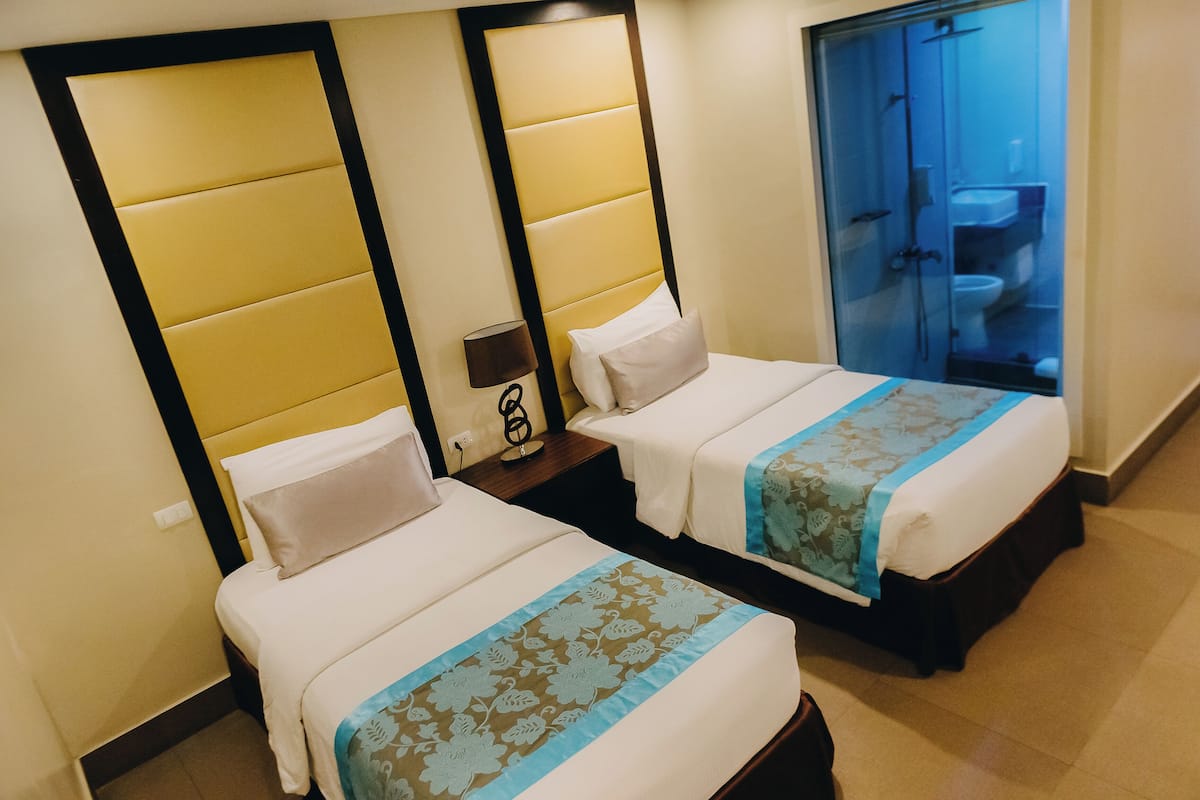 Two twin beds with headboards, a nightstand, a lamp, and a bathroom visible through a glass door.