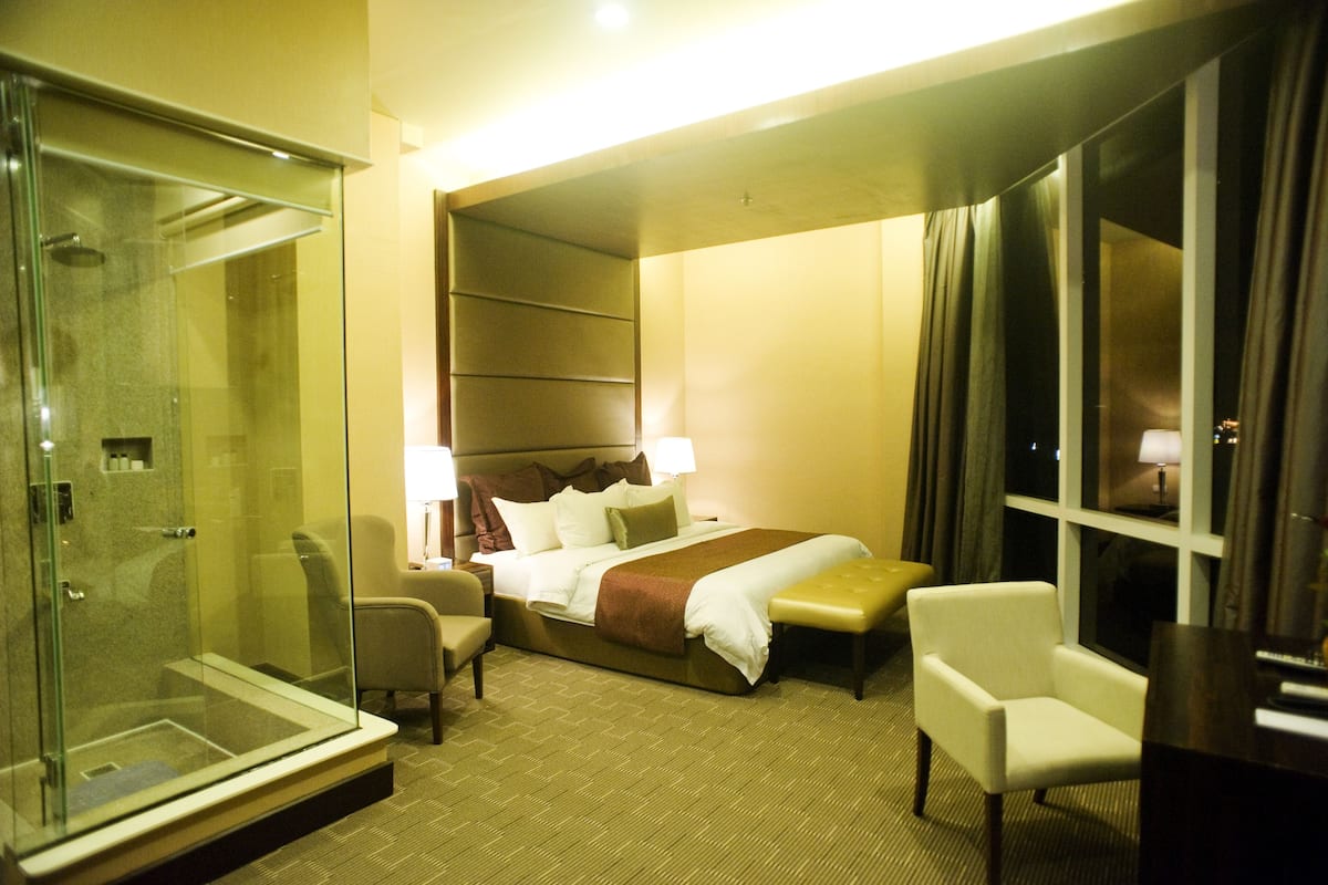 A hotel room with a glass-enclosed shower, a bed with a brown headboard, two bedside tables with lamps, a chair, and a large window with curtains.