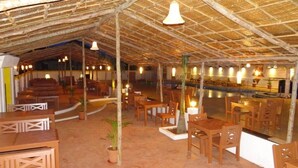 Un restaurant en plein air avec des tables et des chaises en bois, un toit de chaume et des luminaires suspendus.