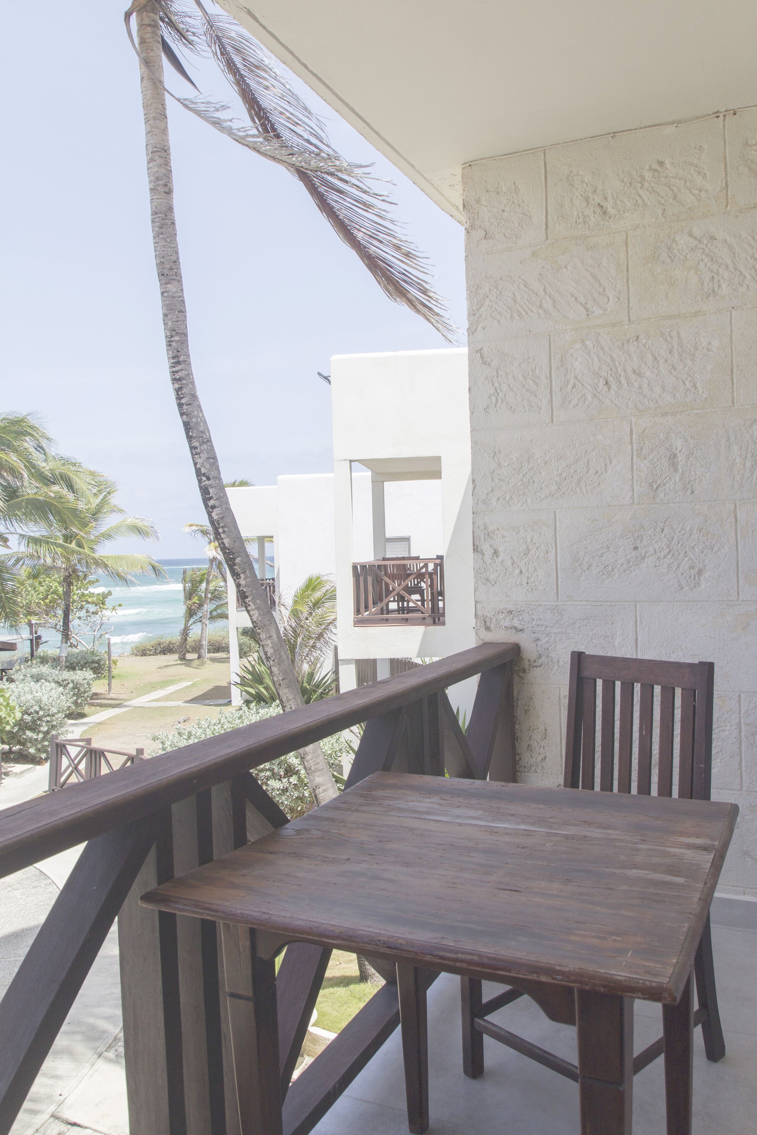 A balcony with a wooden table and chairs, overlooking a beach and palm trees.