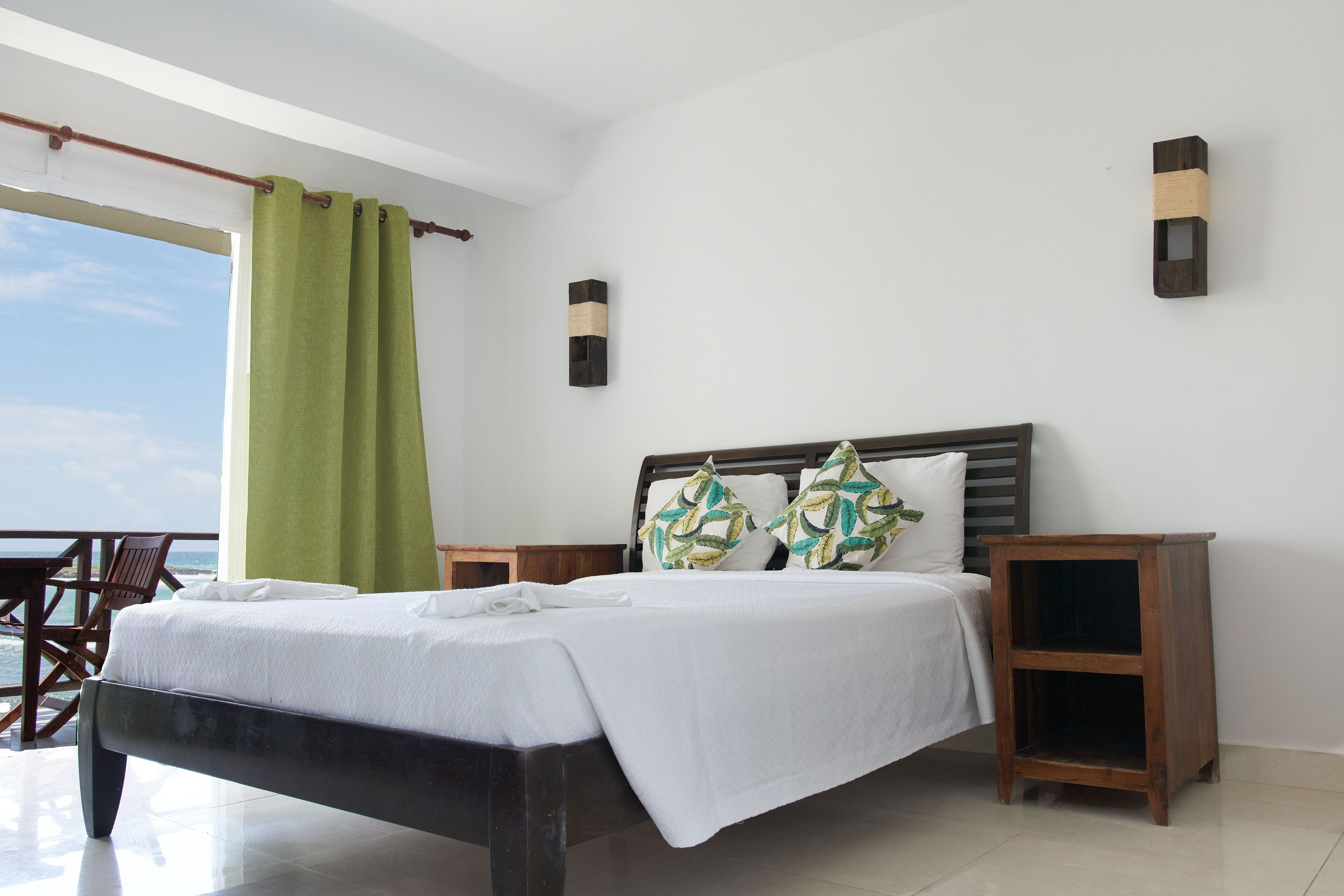 A bedroom with a bed, a nightstand, a wall-mounted light, and a view of the ocean.