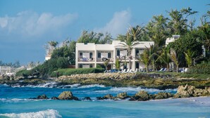 A coastal hotel with multiple balconies overlooking the sea, a rocky shoreline, and palm trees. - Ocean Spray Apartments (Inch Marlowe)
