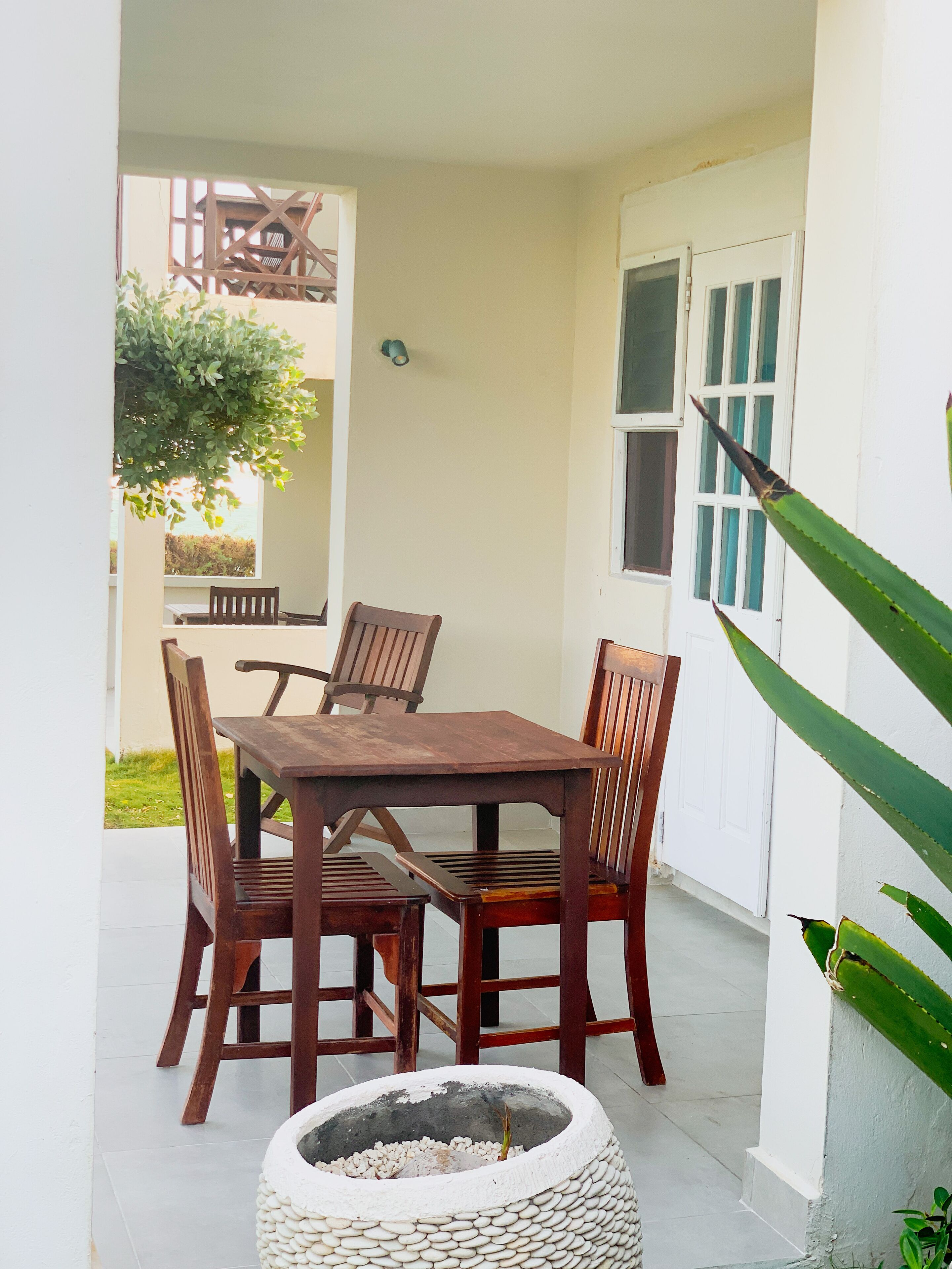 A patio with a wooden table and chairs, a small garden area, and a white wall with a window.