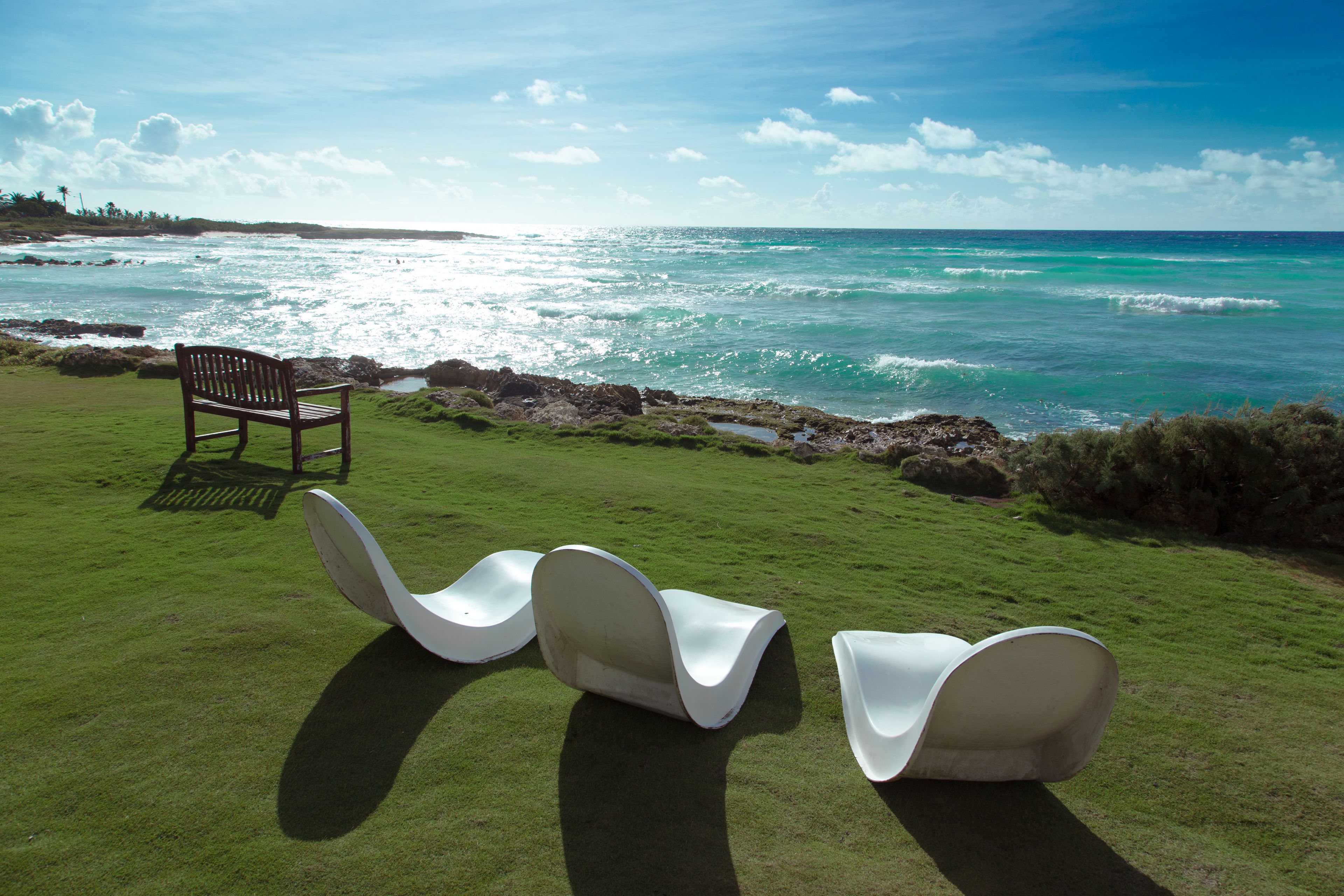 A grassy area with three white lounge chairs and a wooden bench facing a beach with turquoise water.