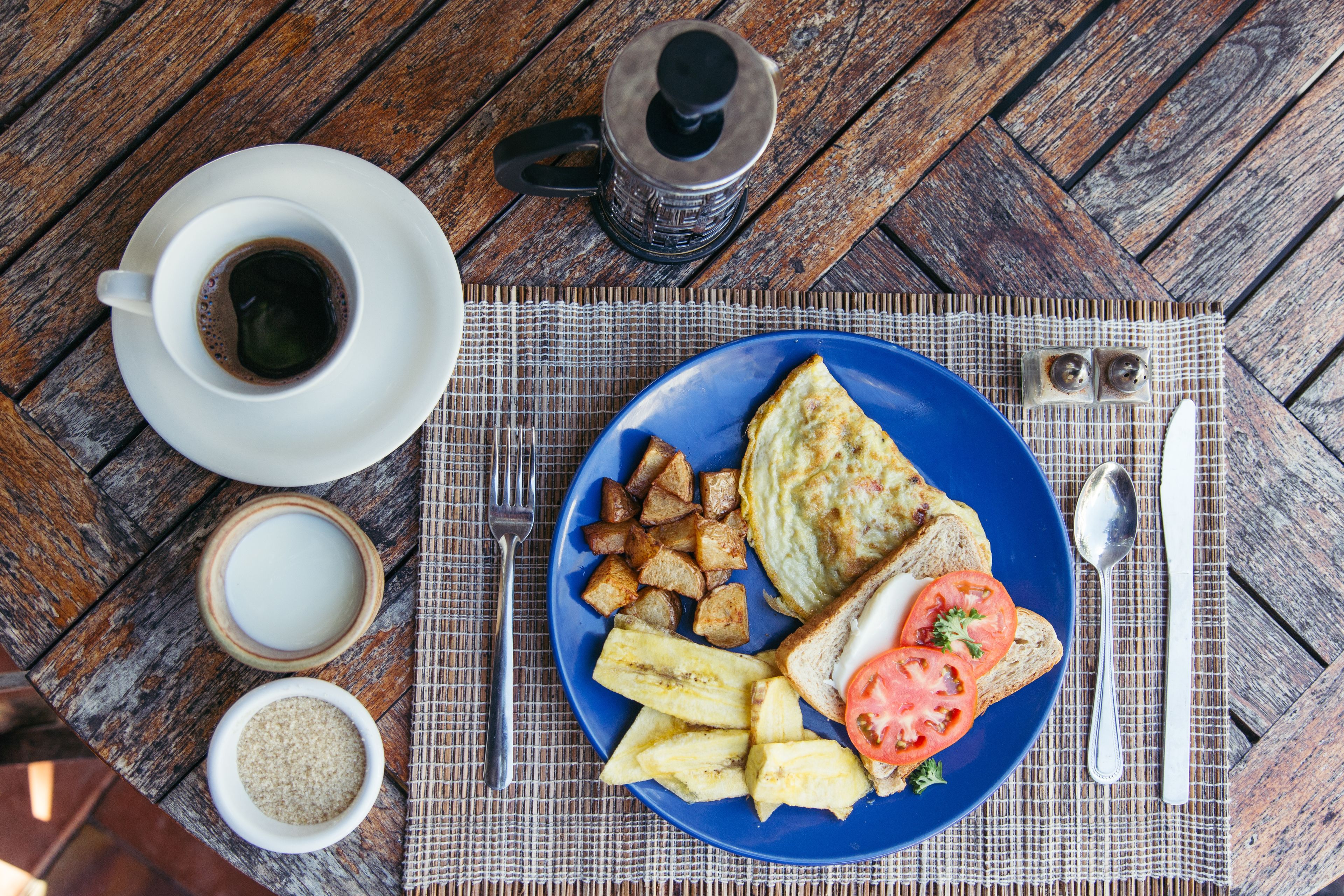 A breakfast plate with toast, eggs, tomatoes, and potatoes, a cup of coffee, and a mug on a wooden table.