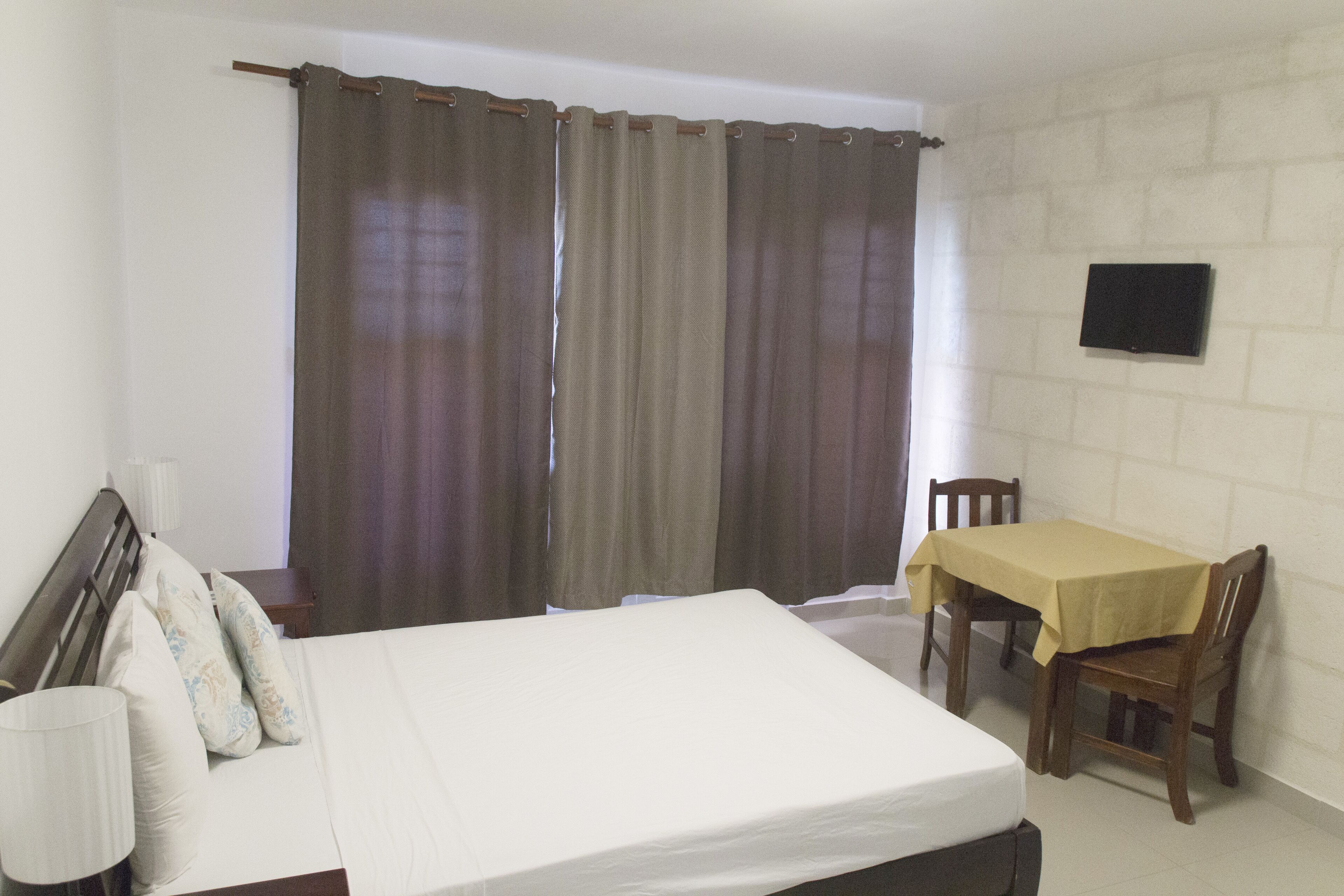 A bedroom with a bed, a table with chairs, a TV on the wall, and curtains.