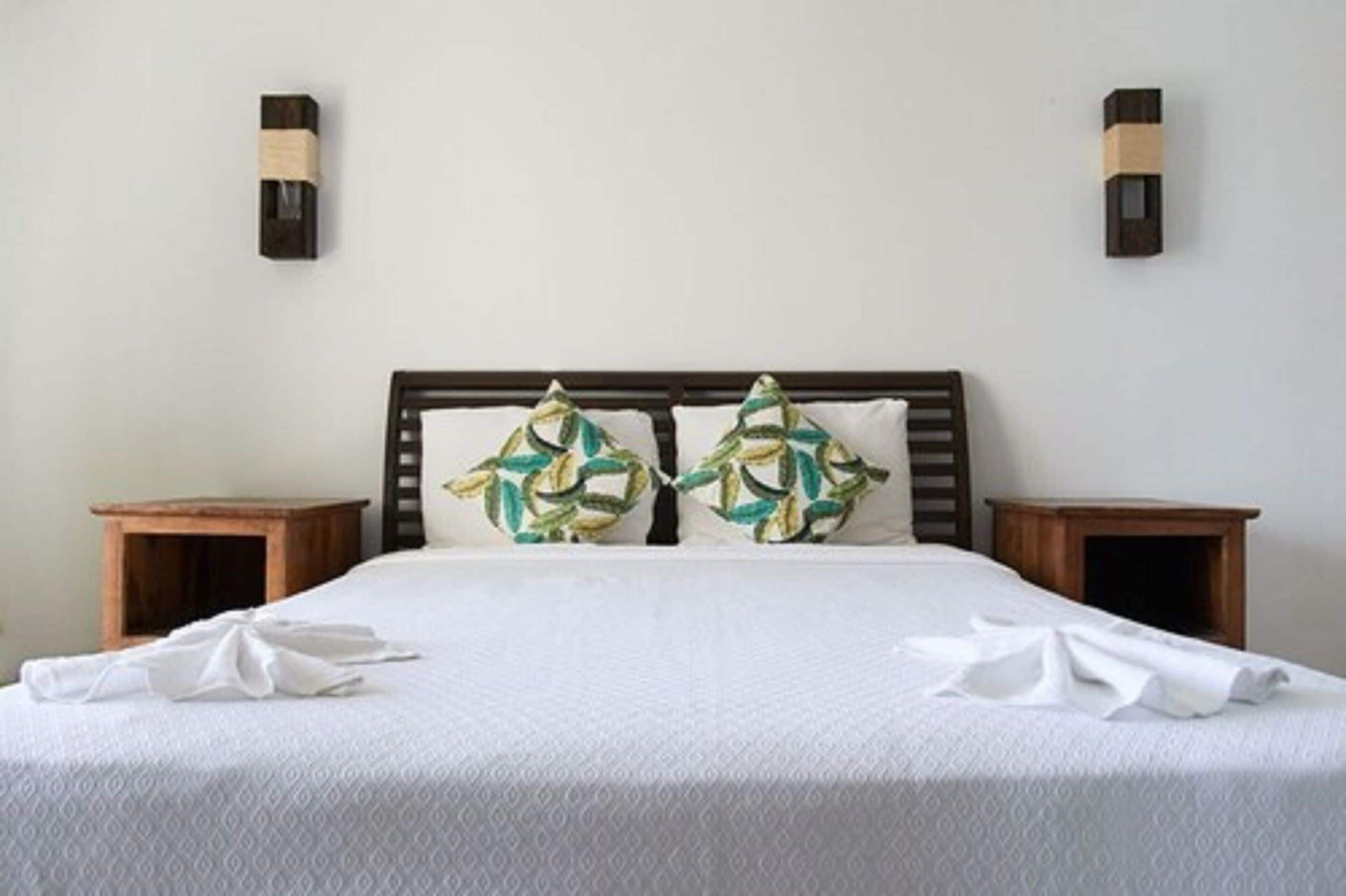Un lit bien fait, recouvert d’une couvre-lit blanche et agrémenté de deux coussins décoratifs, avec deux tables de chevet en bois de chaque côté.