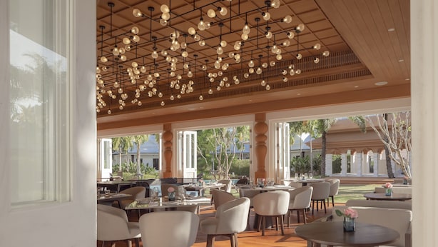 A spacious outdoor dining area with multiple tables and chairs, a wooden ceiling with hanging lights, and large windows offering views of palm trees and a pool. - The Pe La Resort, Phuket (Kamala)