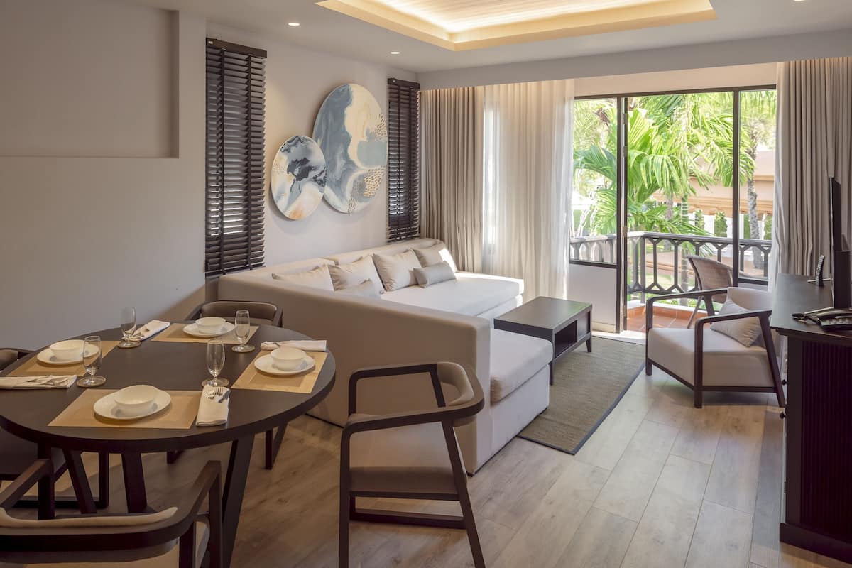 A modern living room with a sofa, dining table, and chairs, a balcony with a view of greenery.