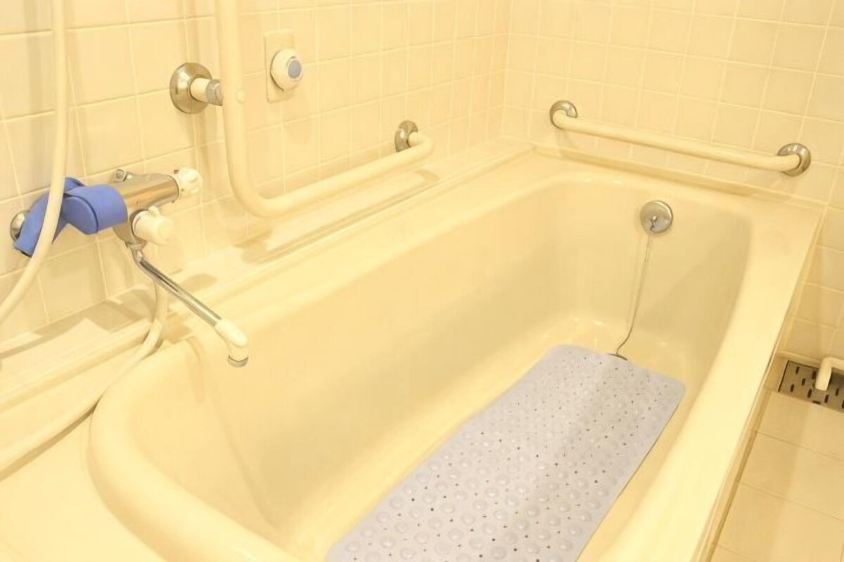 A bathtub with a blue bath mat, grab bars, and a shower control panel.