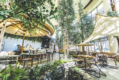 An indoor dining area with tables and chairs, surrounded by greenery and large umbrellas.