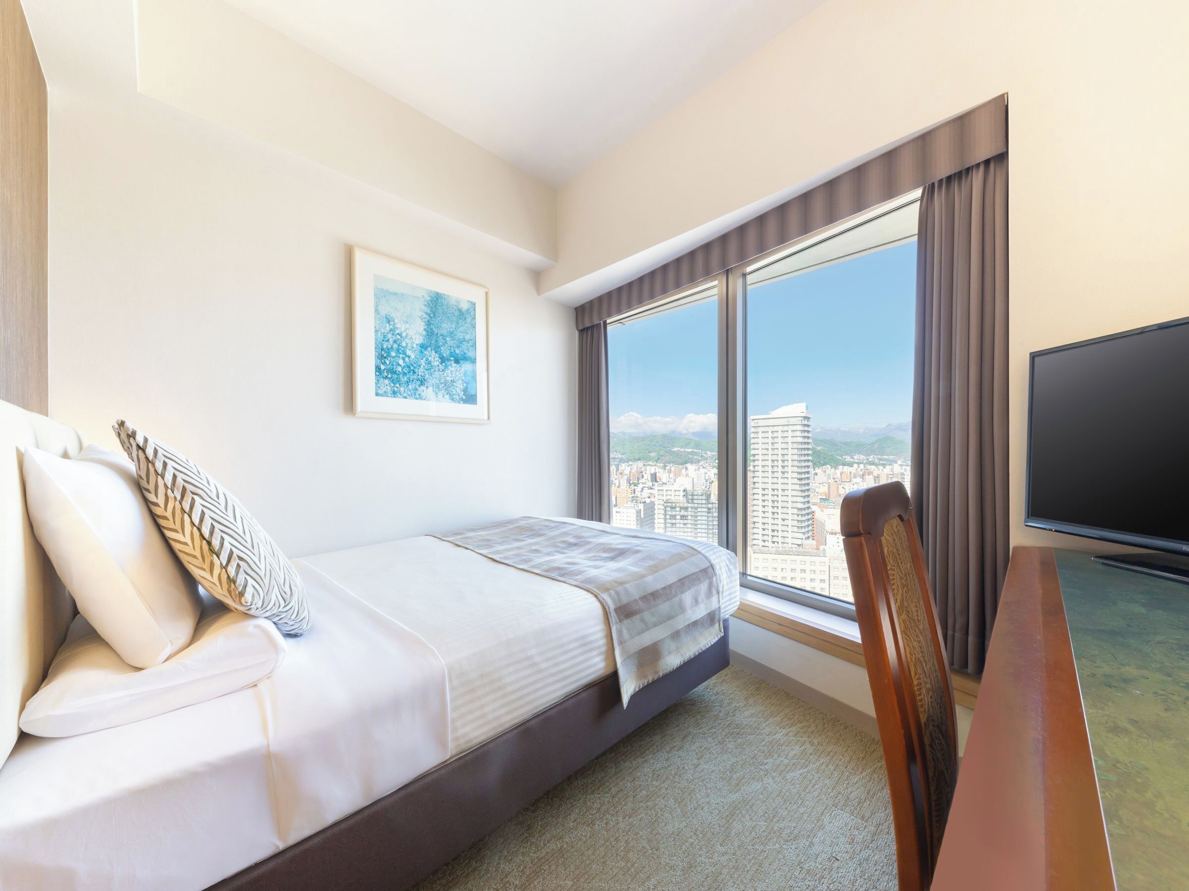 Habitación de hotel con cama, televisor, ventana grande con vista a la ciudad y un cuadro en la pared.