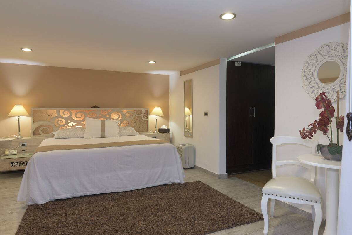 Ein Hotelzimmer mit einem Bett, Nachttischlampen, einem Nachttisch, einem Sessel, einem kleinen Tisch, einem Spiegel und einer Topfpflanze.