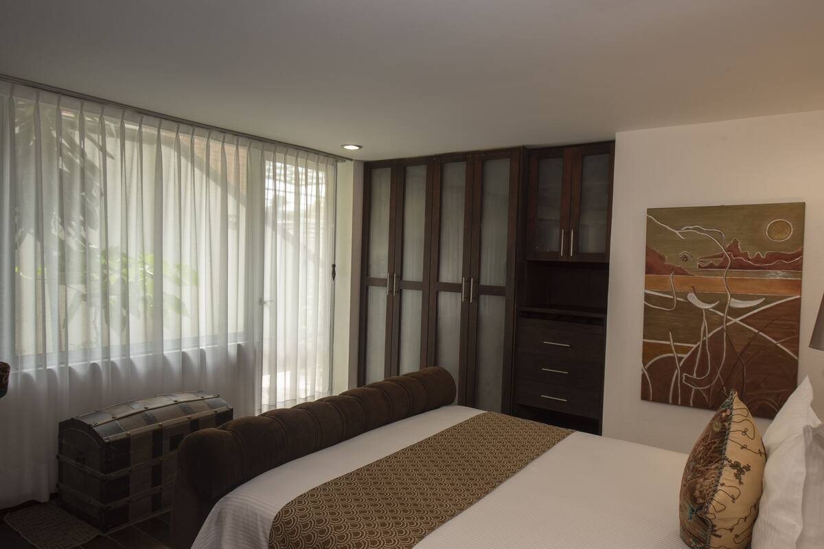 Ein Hotelzimmer mit Bett, einer Kommode, einem Bild an der Wand und einem Fenster mit leichtem Vorhang.