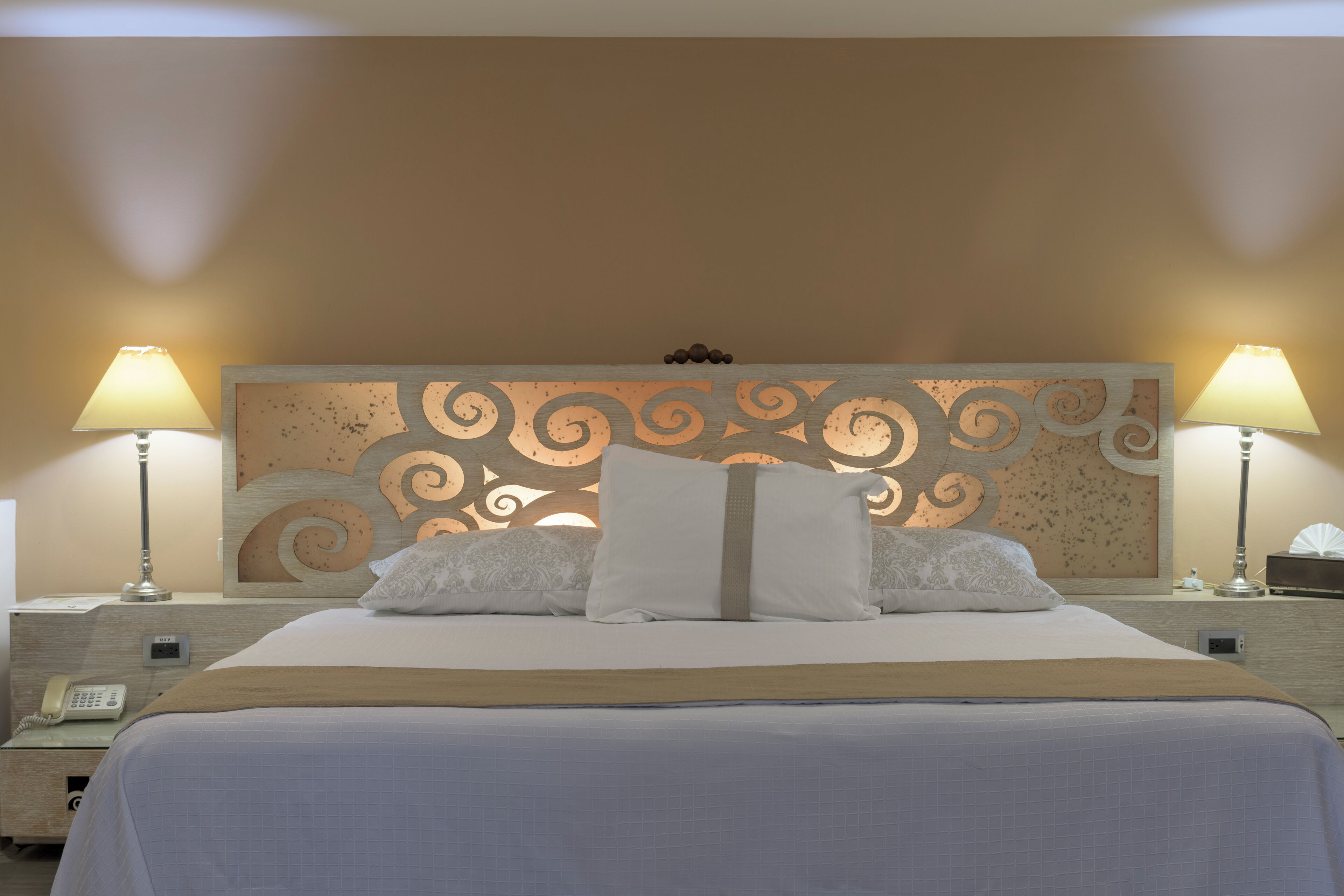 Una cama bien hecha con un cabecero decorativo, dos lámparas de noche y un teléfono junto a la cama.