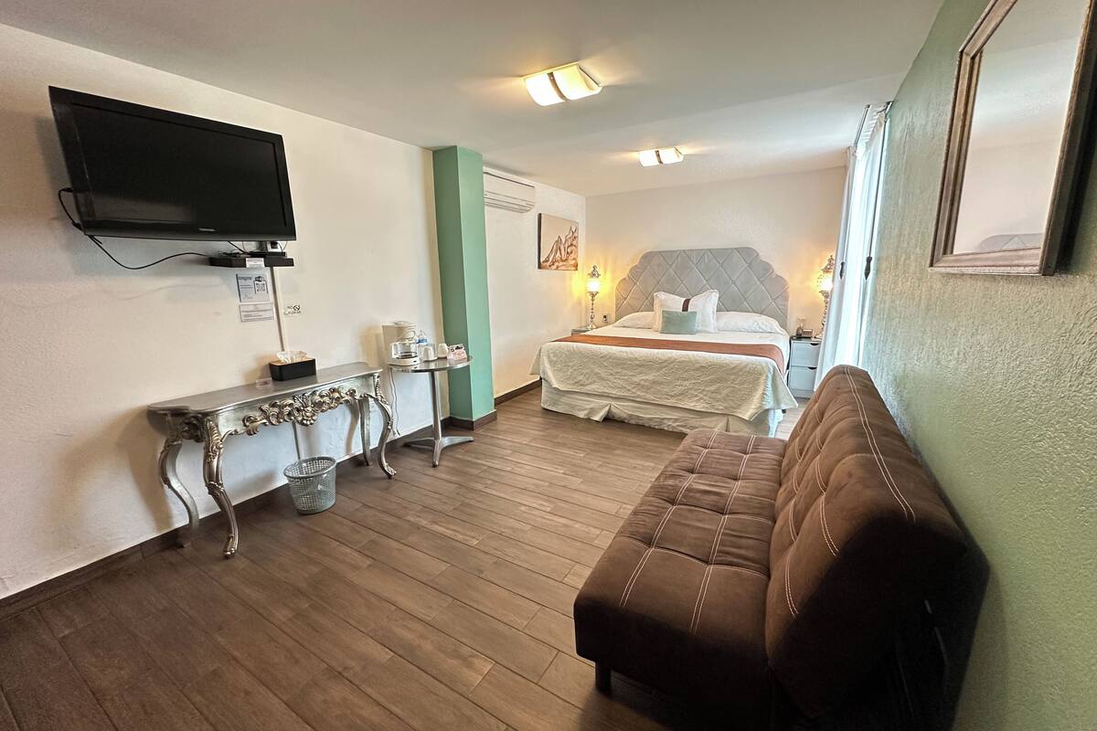 Ein Hotelzimmer mit einem Bett, einem an der Wand montierten Fernseher, einem kleinen Tisch mit einer Lampe und einem braunen Sofa.