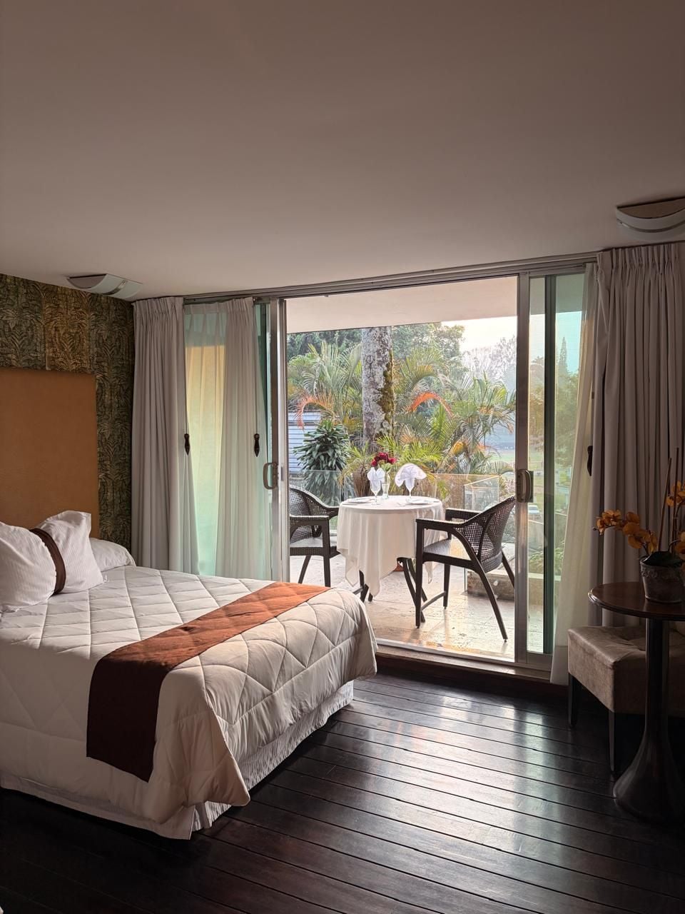 Habitación de hotel con cama, una mesa y sillas, y vistas a un área exterior con plantas a través de un ventanal corredero.