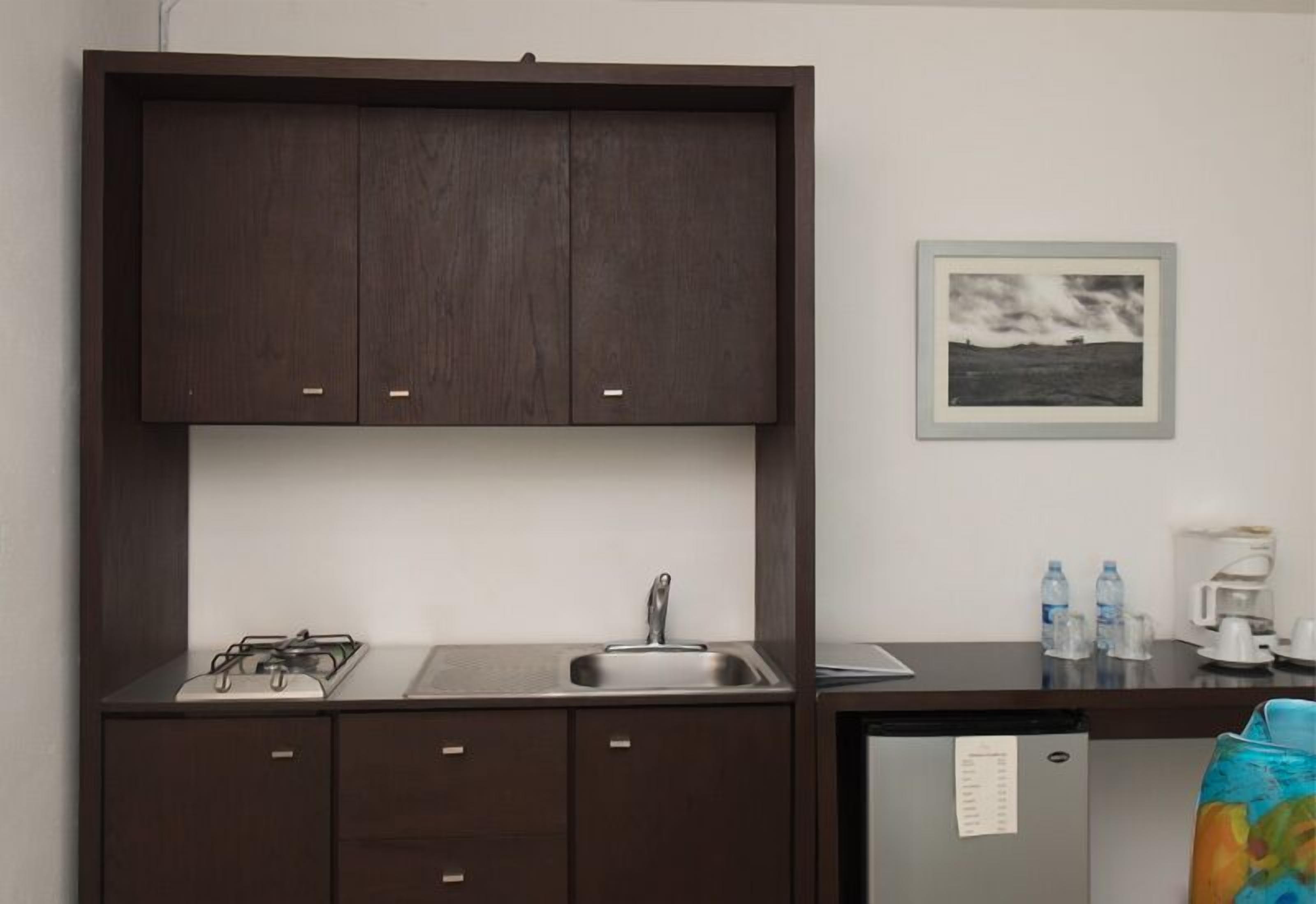 A compact kitchen with a stove, sink, and a refrigerator. There is a framed picture on the wall and a small table with a coffee maker and water bottles.