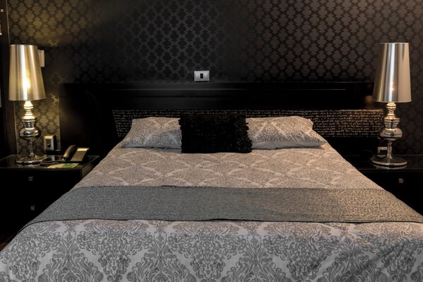 A neatly made bed with a patterned bedspread and pillows, two bedside lamps, a nightstand with a phone and a lamp, and a dark headboard with a light switch. - Colombe Hotel Boutique (Xalapa)