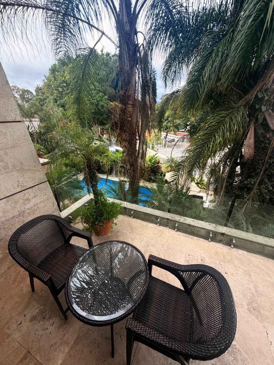 A balcony with two wicker chairs and a glass table, overlooking a pool and lush greenery.