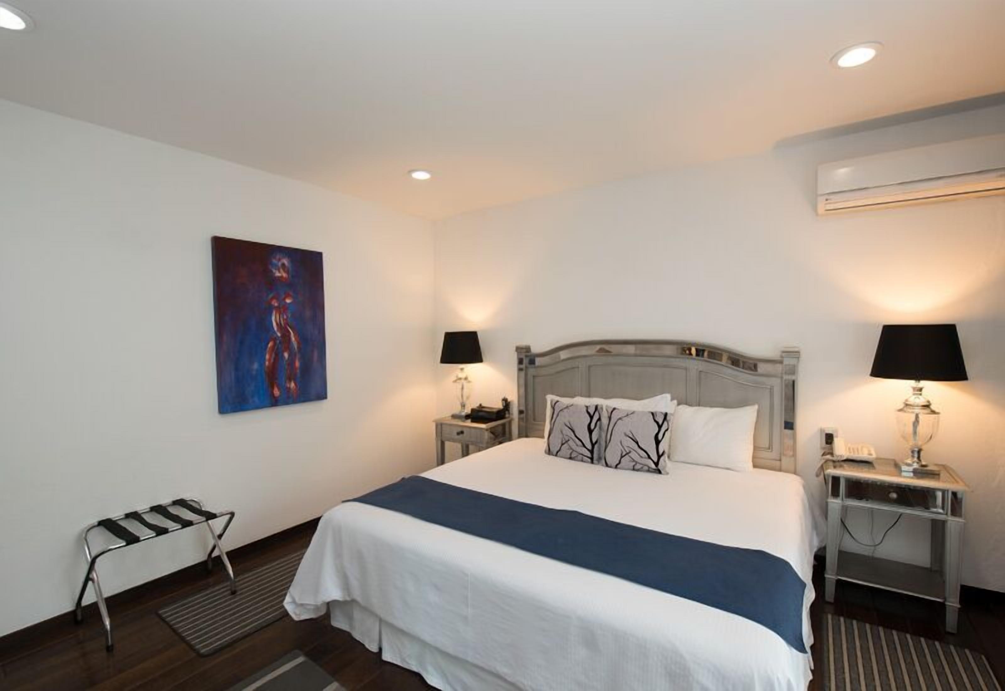 A hotel room with a large bed, two bedside tables with lamps, a bench, a painting on the wall, and an air conditioning unit.