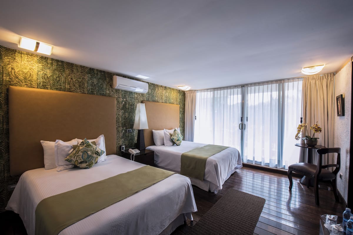 Ein Hotelzimmer mit zwei Betten, einem Schreibtisch, einem Sessel, einem Fenster mit Vorhängen und einer Klimaanlage.