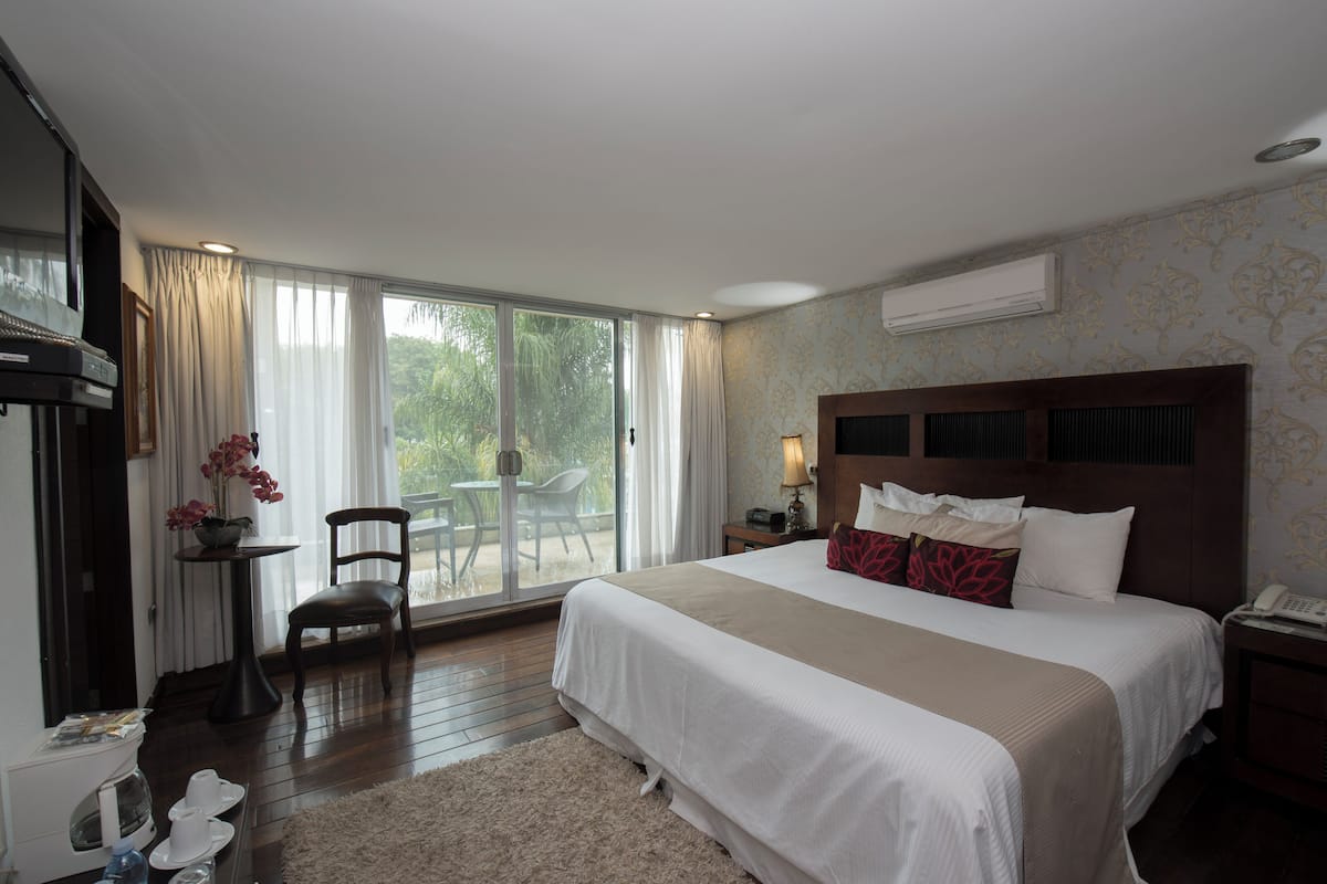 Ein Hotelzimmer mit einem großen Bett, einem Sessel, einem kleinen Tisch mit einer Lampe und Blick ins Freie durch eine Schiebetür.