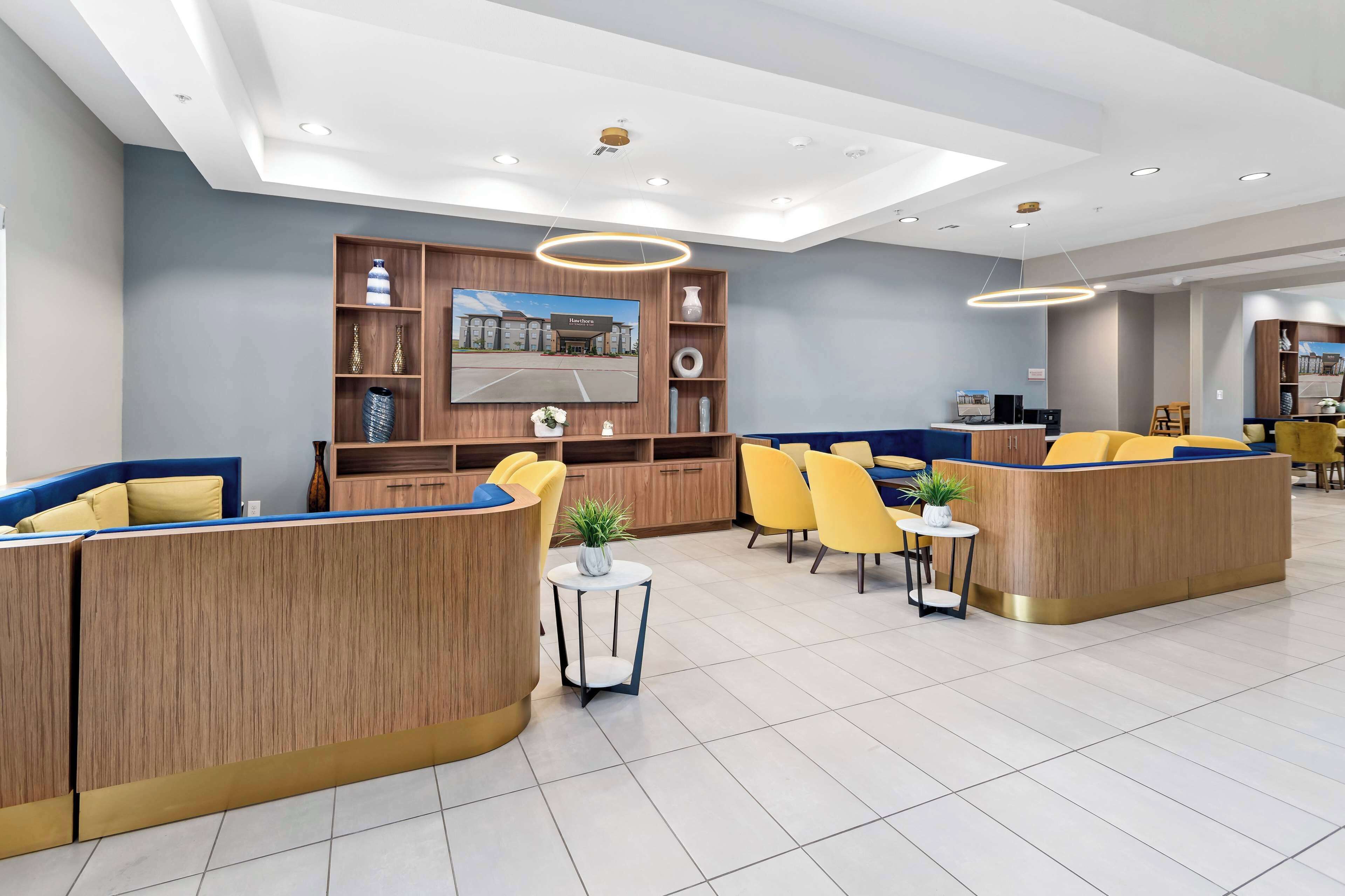 A modern hotel lobby with wooden reception desks, yellow chairs, and a large TV screen displaying a hotel exterior.