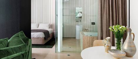 Una habitación de hotel moderna con una cama, cabina de ducha de vidrio, baño con lavamanos y espejo, y un comedor con mesa dispuesta para dos.