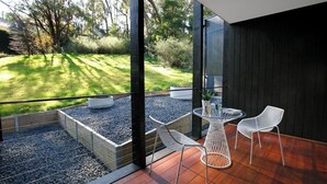A modern outdoor patio with a wooden deck, a glass table, and two white chairs. - Hepburn at Hepburn (Hepburn Springs)
