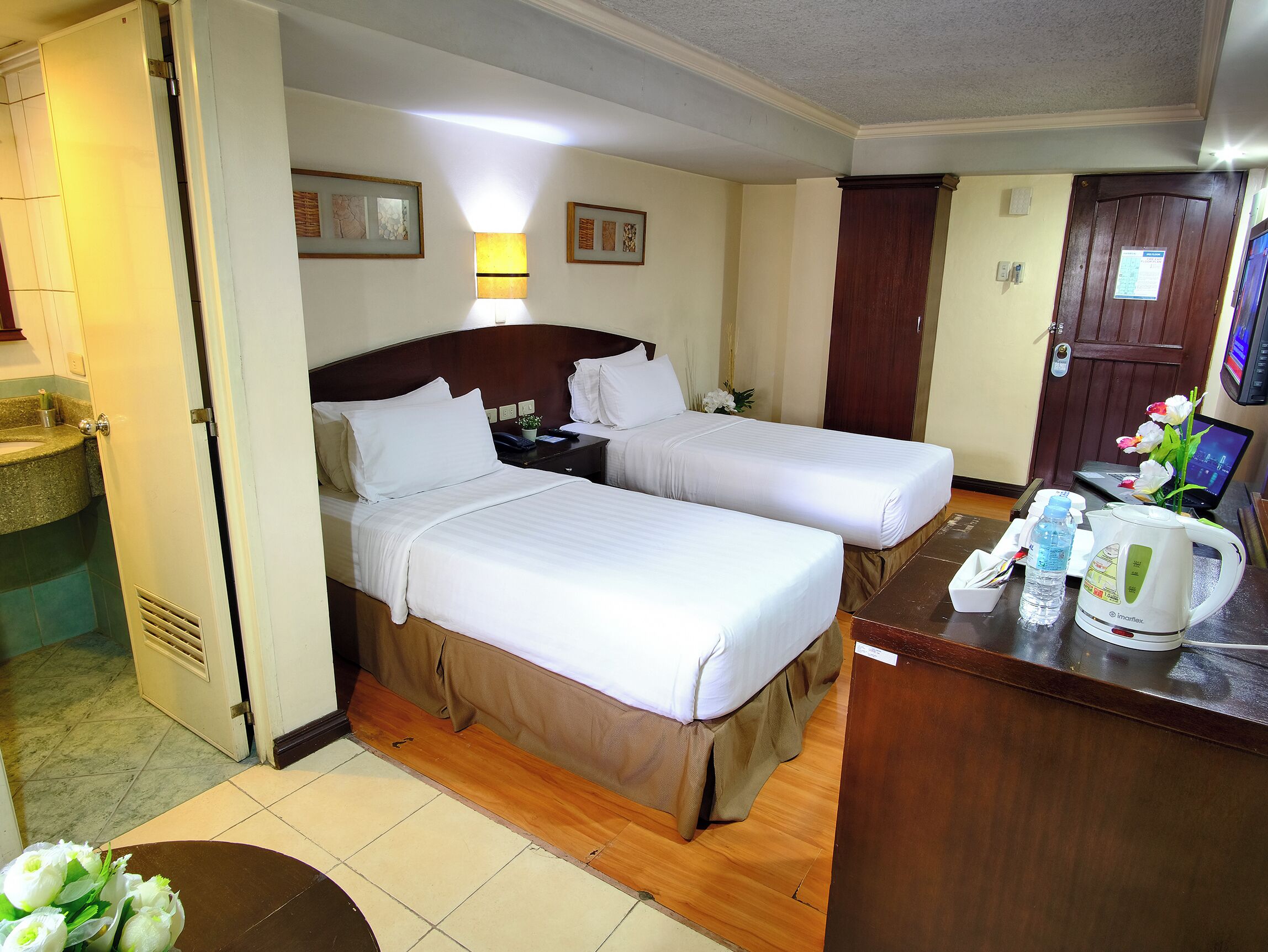 A hotel room with two beds, a desk with a laptop, a coffee maker, and a small table with a vase of flowers.