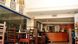 Um hall de hotel com balcão de recepção, cadeiras de madeira, área de refeições e um padrão decorativo no chão.