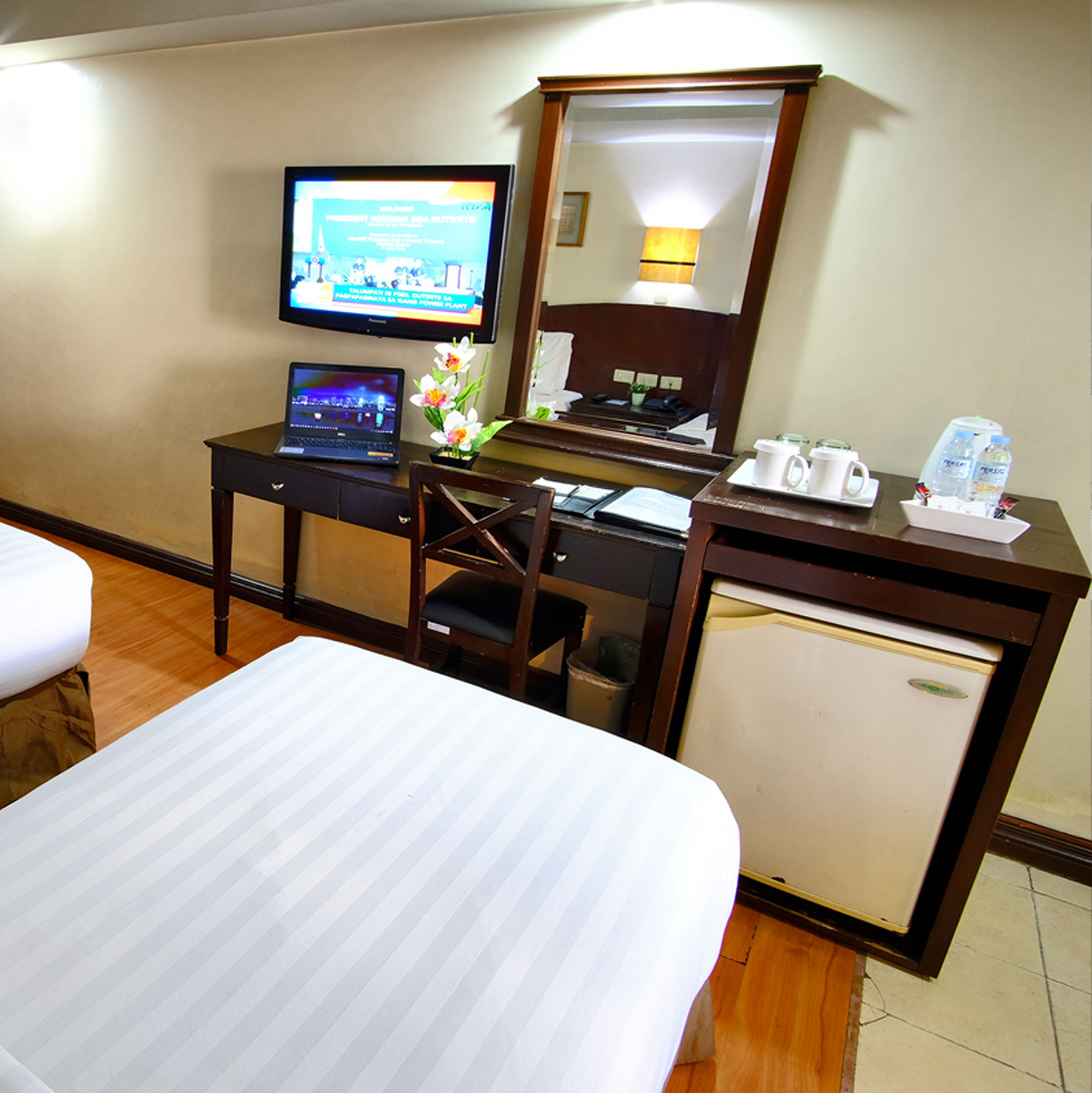 Quarto de hotel com uma cama, uma escrivaninha com um laptop, uma televisão, um frigorífico e uma pequena mesa com uma chávena e garrafas de água.