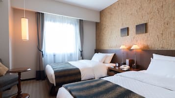 A hotel room with two beds, a small table, a chair, a nightstand with a lamp, and a window with curtains.