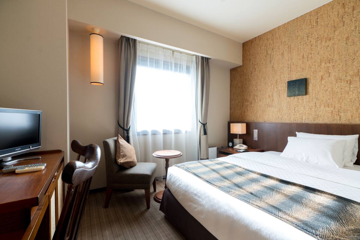 A hotel room with a bed, a desk with a TV, a chair, a small table, and a window with curtains.