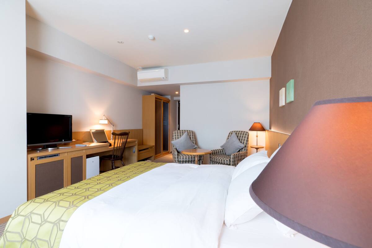 A hotel room with a bed, a desk with a television, two armchairs, and a lamp.
