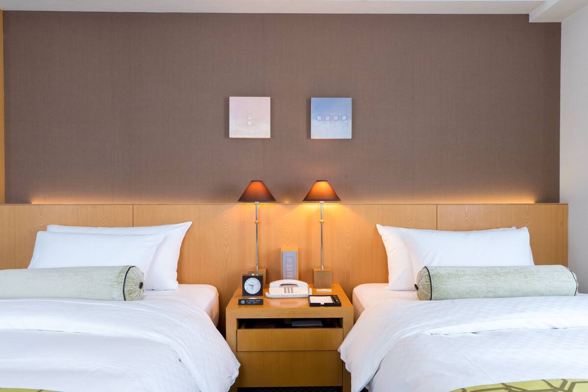 A hotel room with two beds, a wooden headboard, a nightstand with a lamp, a clock, and a telephone.