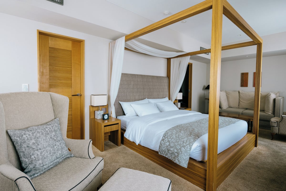Un lit à baldaquin avec un cadre en bois, un fauteuil beige agrémenté d’un coussin à motifs, une table de chevet avec une lampe et une porte en bois.
