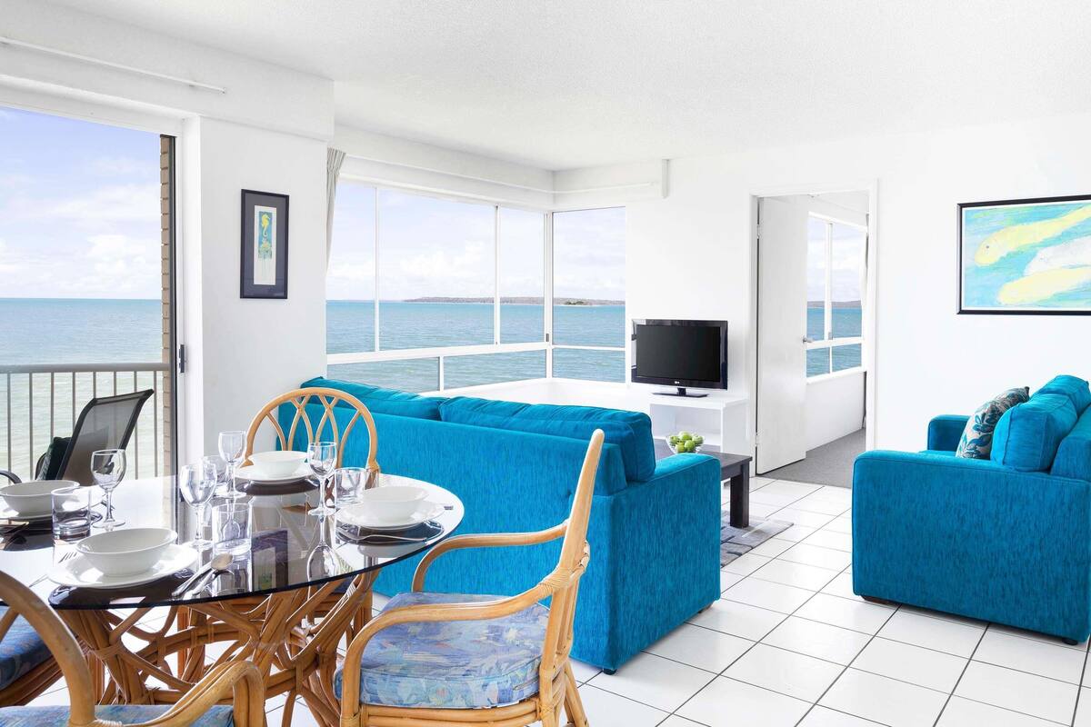 Ein heller Raum mit gedecktem Esstisch, blauen Sofas, einem Fernseher und Meerblick.