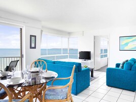 Habitación luminosa con mesa de comedor, sofás azules, televisor y vista al mar.