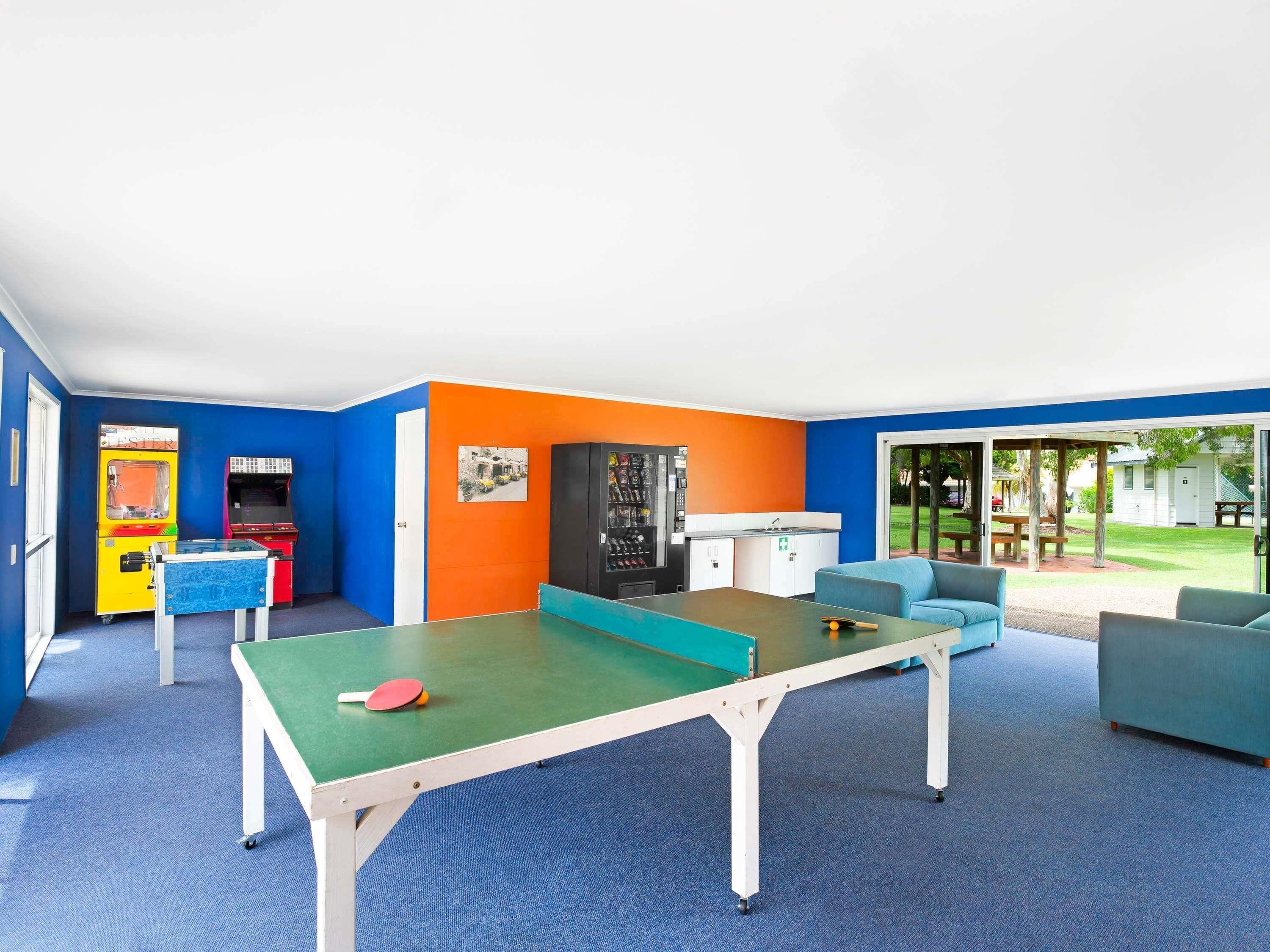 A room with a ping pong table, arcade games, a kitchenette, and a view of a grassy area.