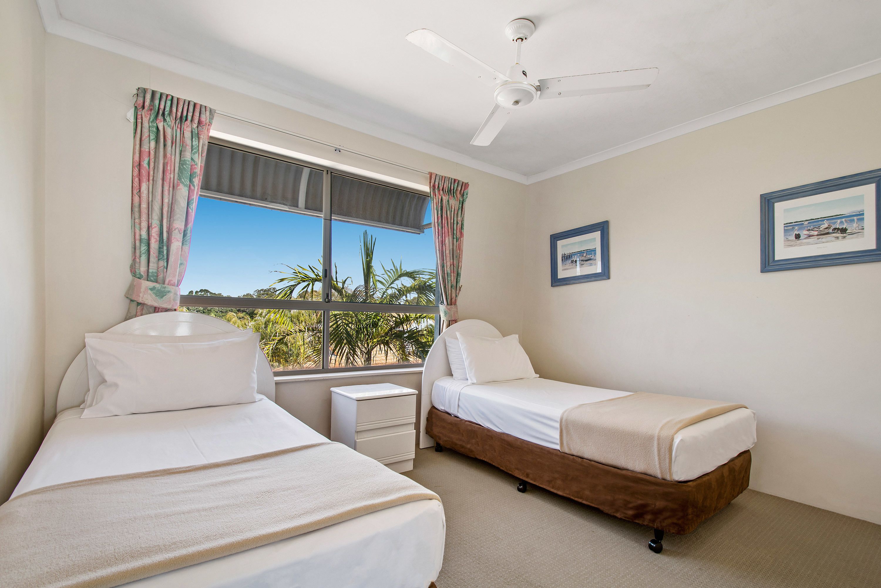A bedroom with two beds, a ceiling fan, a window with curtains, and a small bedside table.