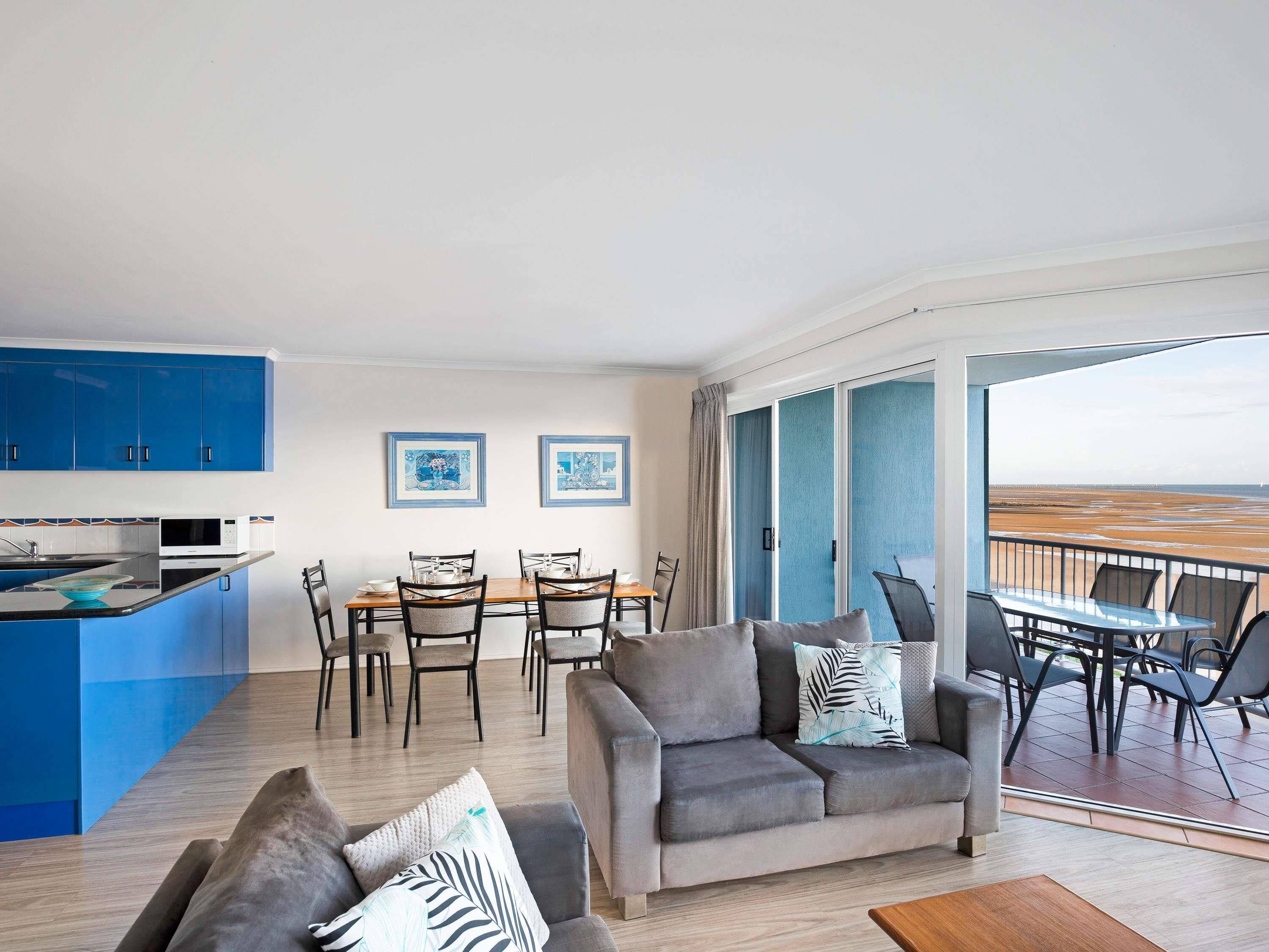 A modern living room with a sofa, dining table, and chairs, a kitchen area, and a balcony with a view.