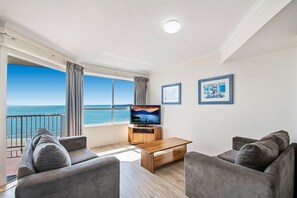 A modern living room with a large window overlooking the sea, a wooden coffee table, and two grey sofas. - BreakFree Great Sandy Straits (Urangan)
