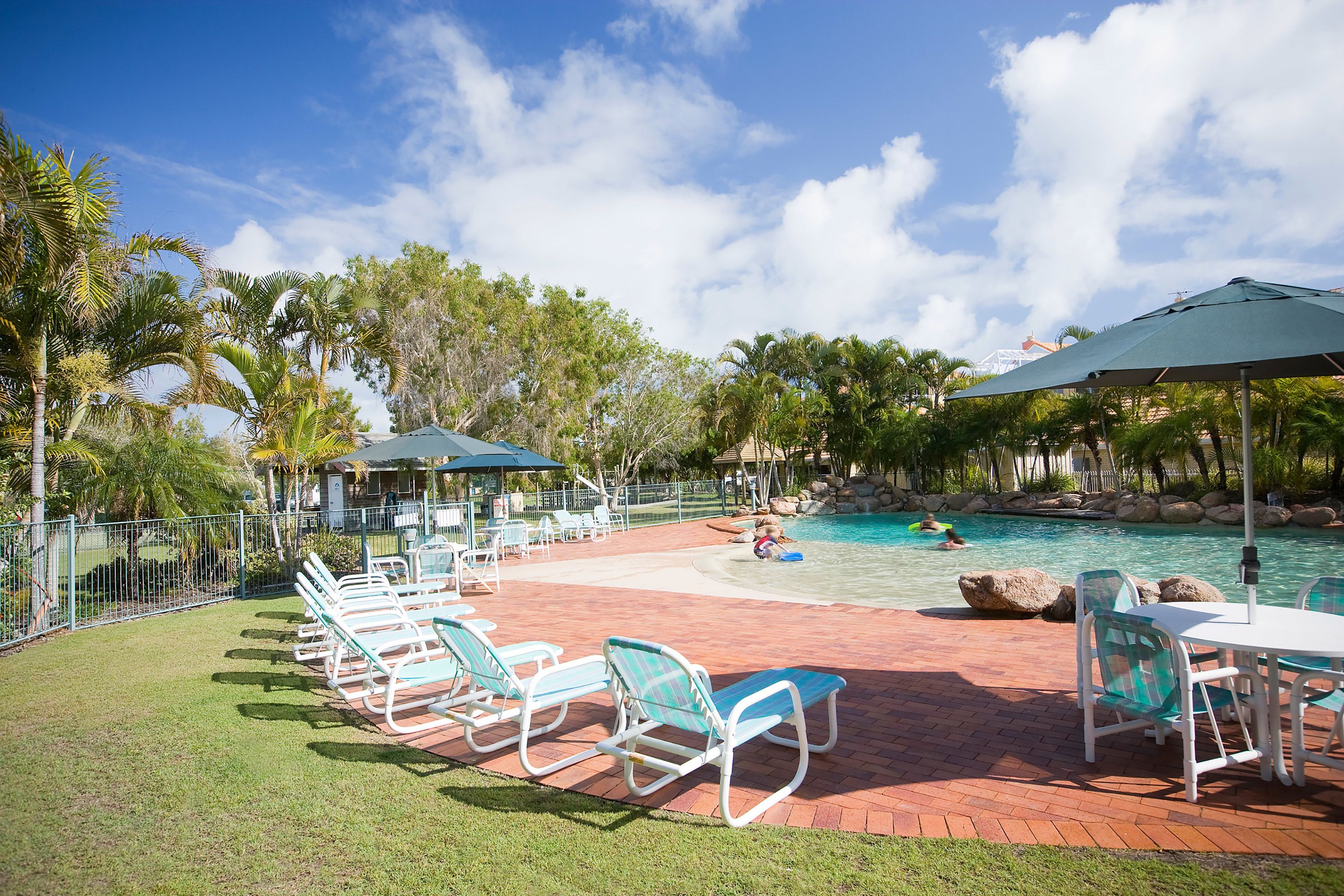 A pool area with lounge chairs and umbrellas, surrounded by palm trees and a clear sky.