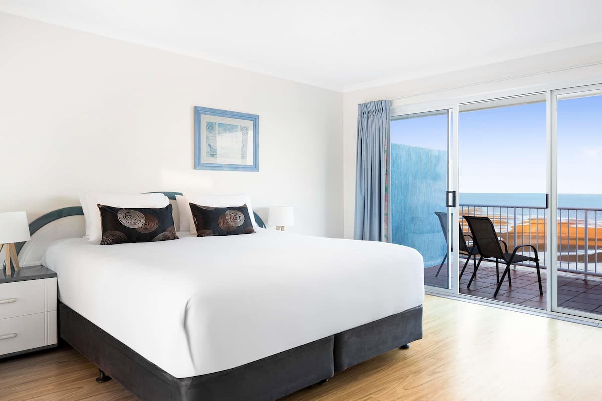 Ein Hotelzimmer mit einem großen Bett, einem Nachttisch, einem Sessel und einem Balkon mit Blick auf den Strand.